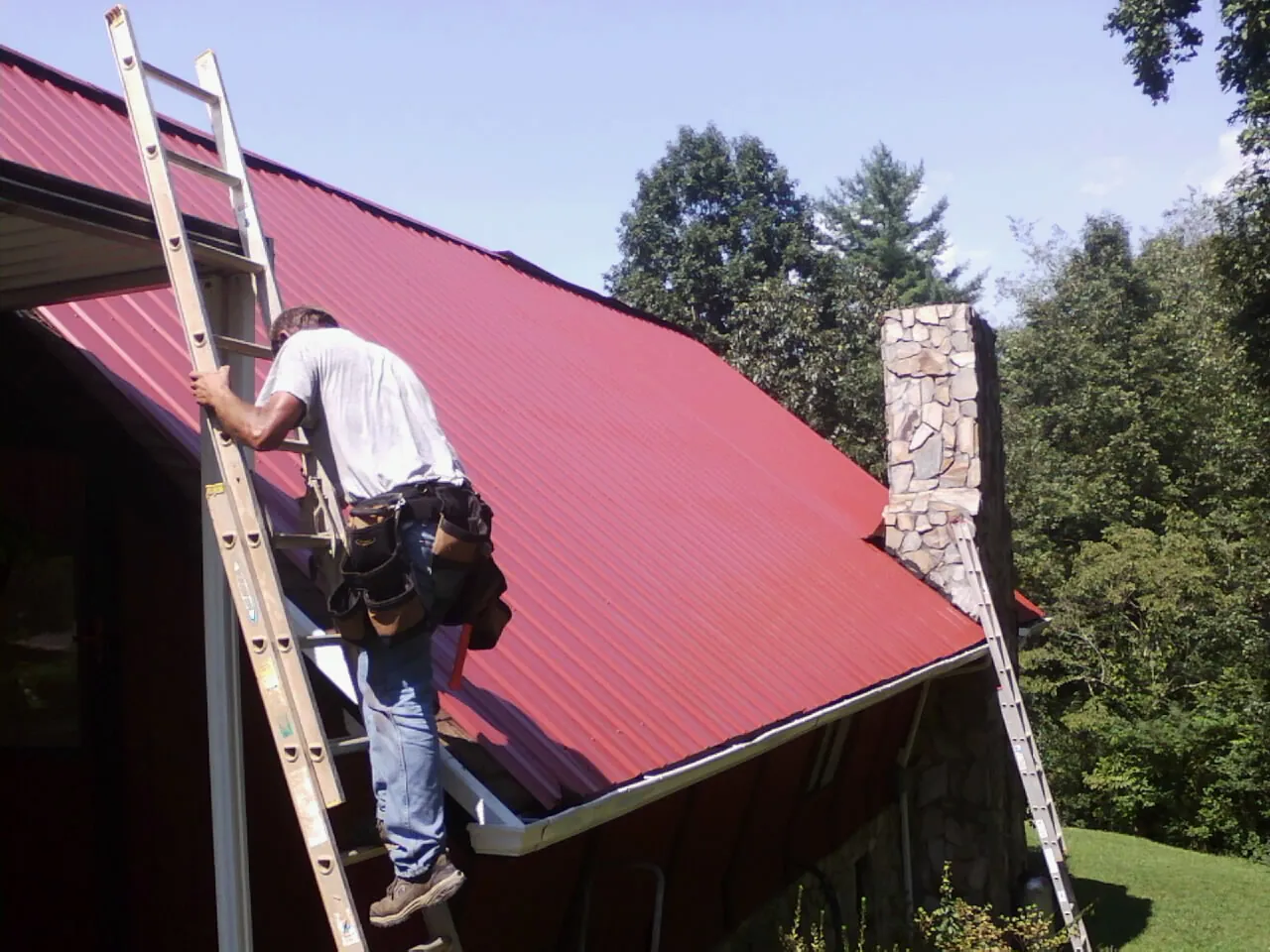 Roofer on a ladder beside a red metal roof and stone chimney — Soffit & Fascia Repair by Roofing Brothers in Horse Creek